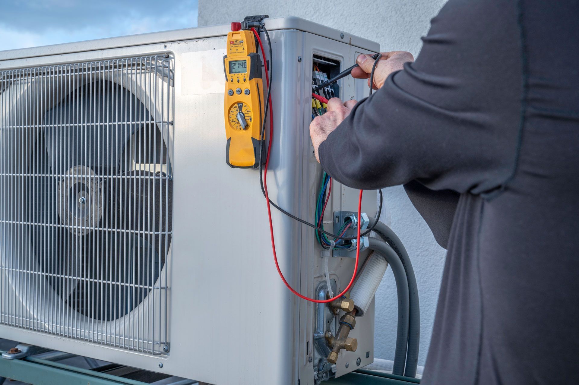 Technician using a meter to service an outdoor HVAC unit with exposed wiring and gauges.