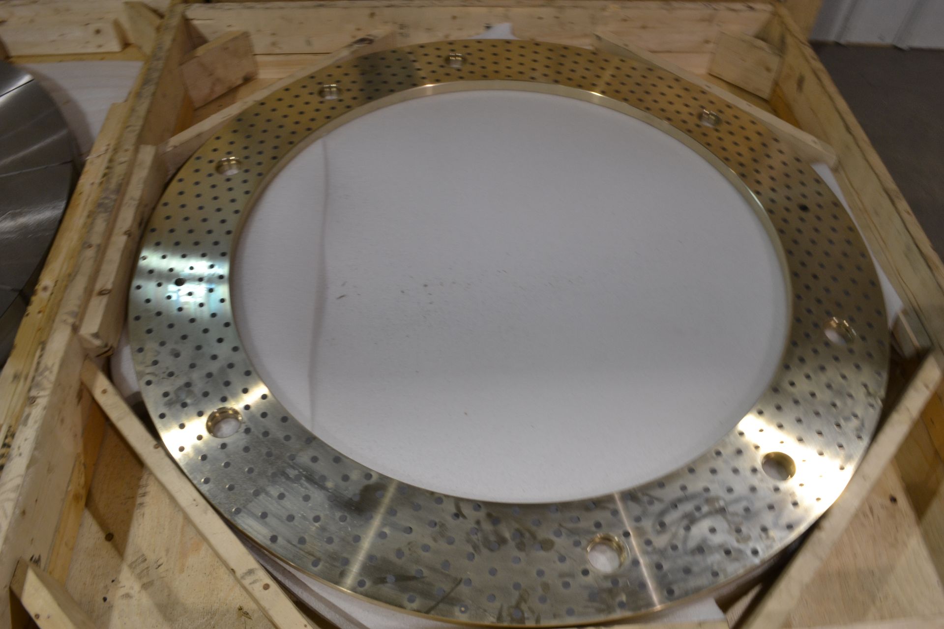 Large golden metal disc with numerous holes and a central white area, in a wooden crate.
