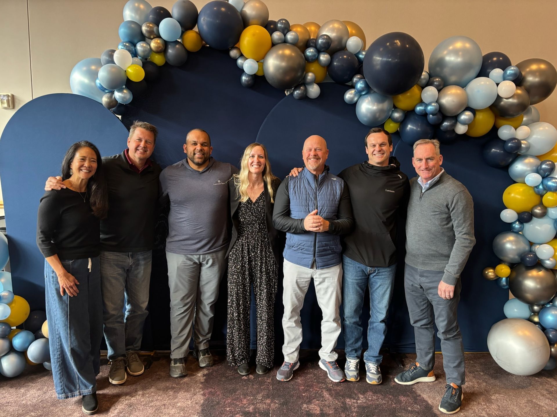 Seven smiling people stand in a line before a blue backdrop decorated with an arch of blue, gold, and silver balloons.