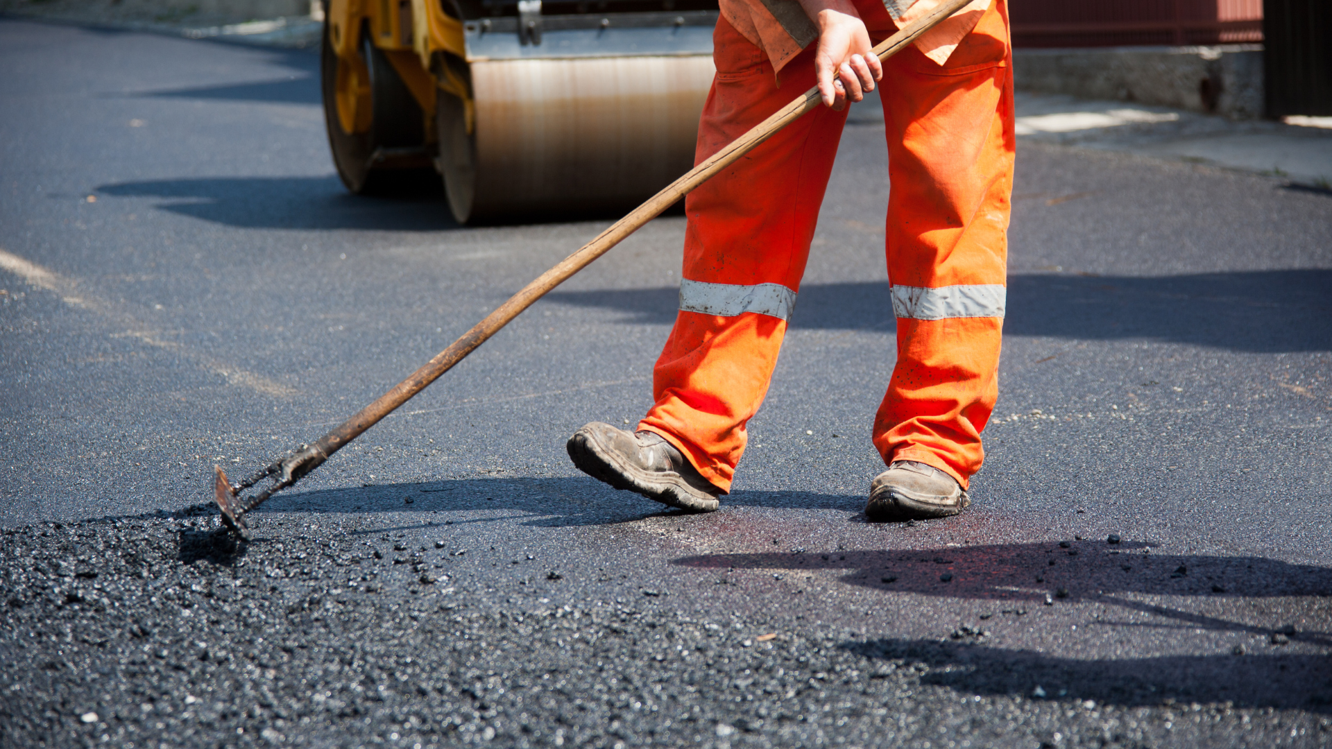 A man in orange pants is spreading asphalt on a road.