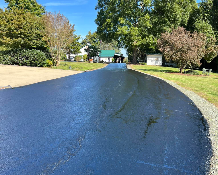 A black asphalt driveway leading to a house surrounded by trees.