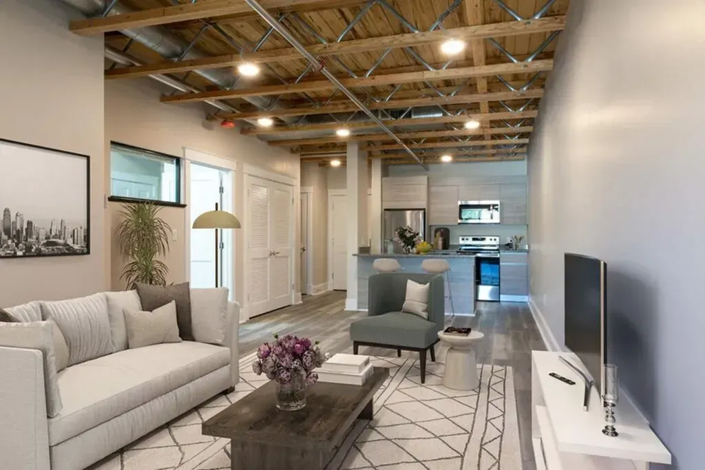 Living room with exposed wood ceiling, grey walls, couch, armchair, TV, and kitchen in the background.
