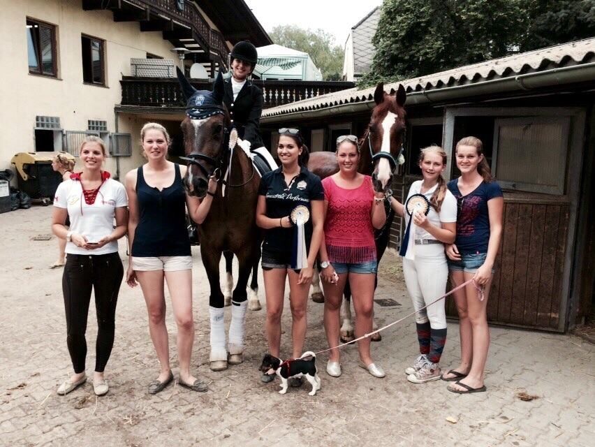 Eine Gruppe von Menschen posiert mit Pferden vor einem Stall. Auf einem Pferd sitzt ein Reiter; einige halten die Zügel, andere stehen daneben.