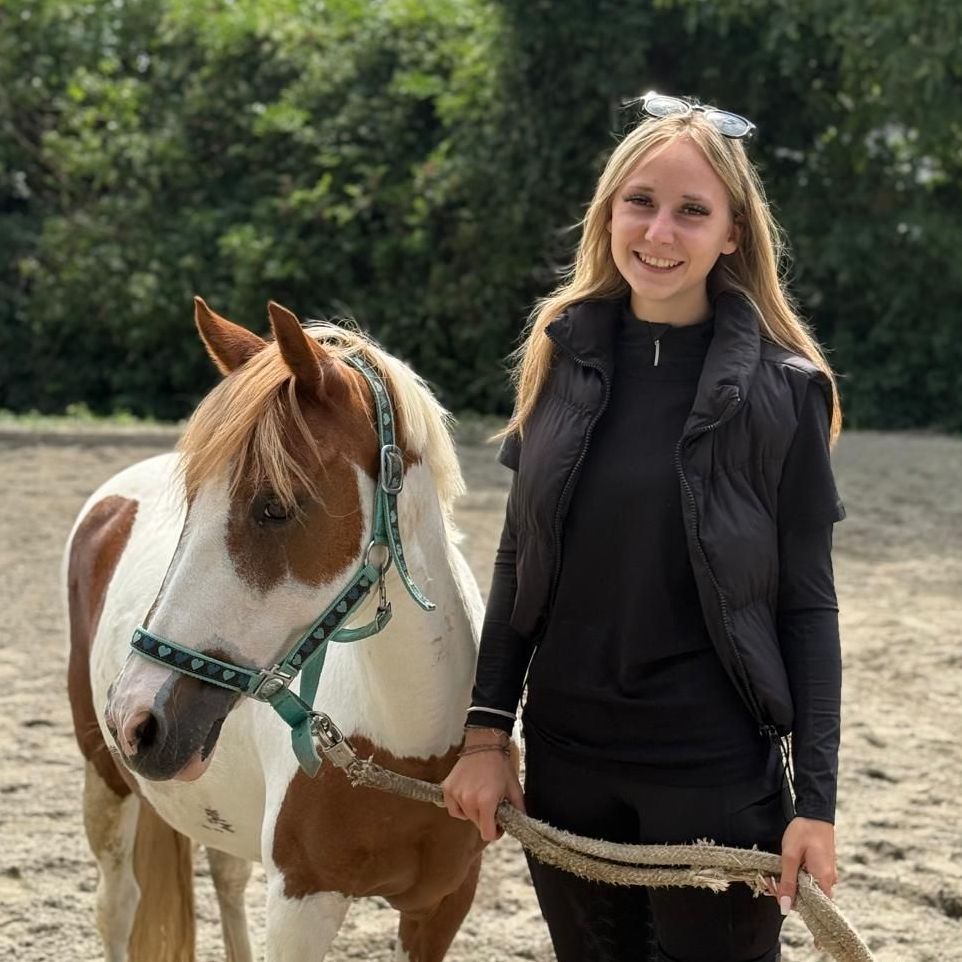 Eine junge Frau lächelt neben einem Paint Horse auf einer Reitbahn. Das Pferd hat braune und weiße Abzeichen; die Frau trägt Schwarz.