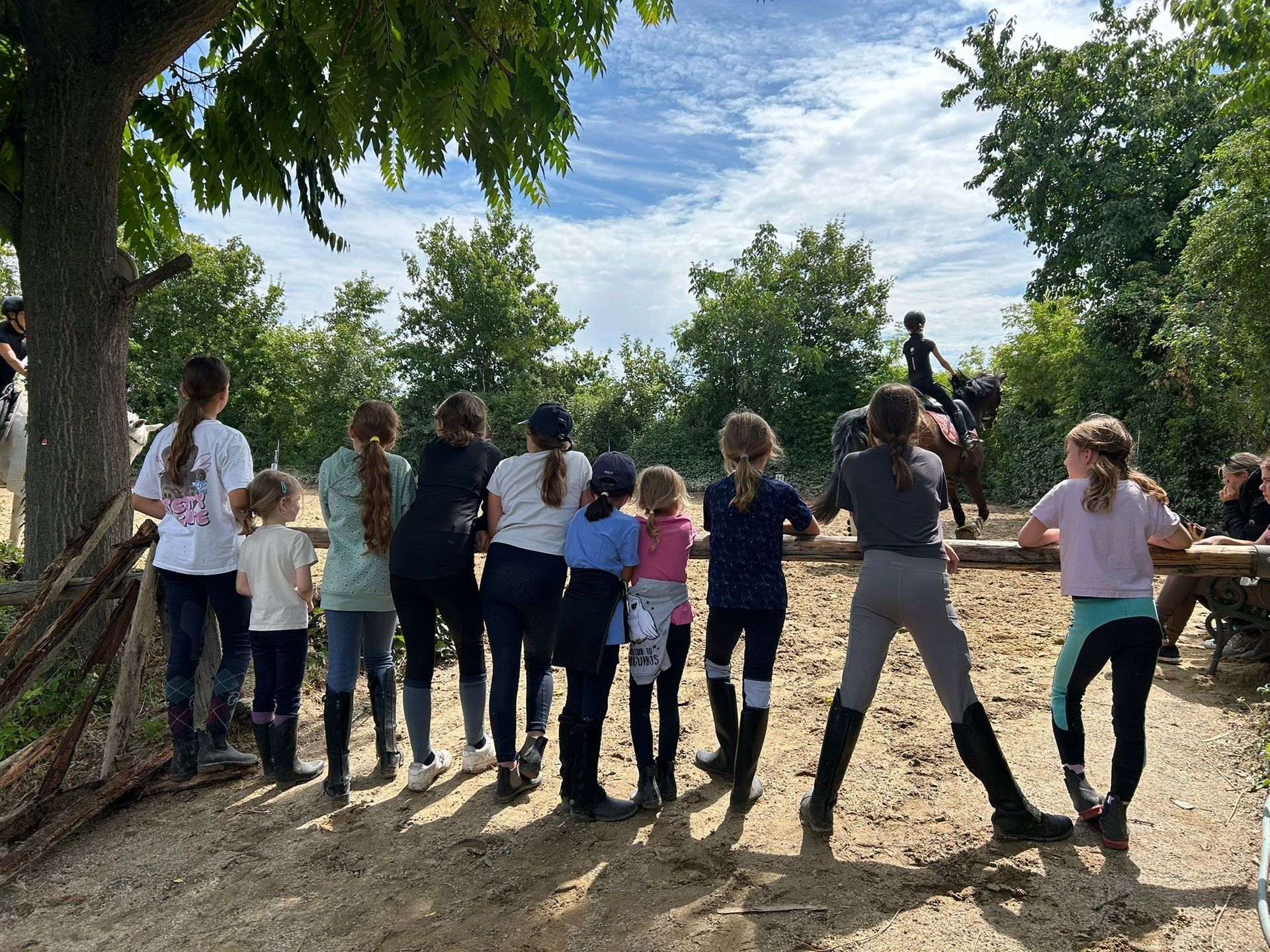 Kinder beobachten einen Reiter in einer Außenarena. Sie stehen hinter einem Holzzaun unter einem bewölkten Himmel.