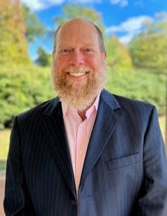 A man with a beard is wearing a suit and a pink shirt.
