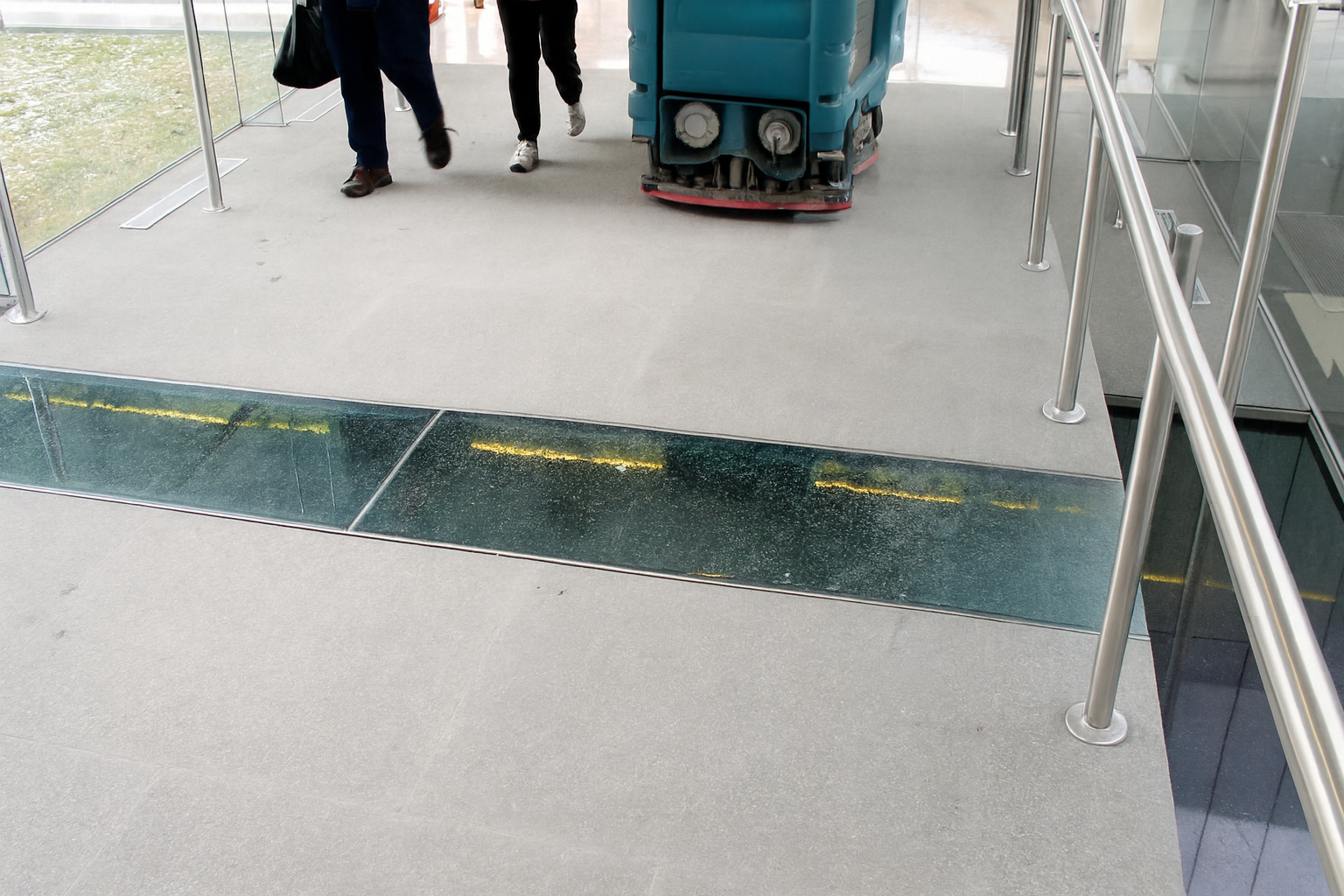 People walk past a floor cleaning machine and a glass section on a sidewalk, near a railing.