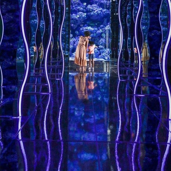 People in a mirrored room with blue lighting, creating a psychedelic effect. Blue glass floor