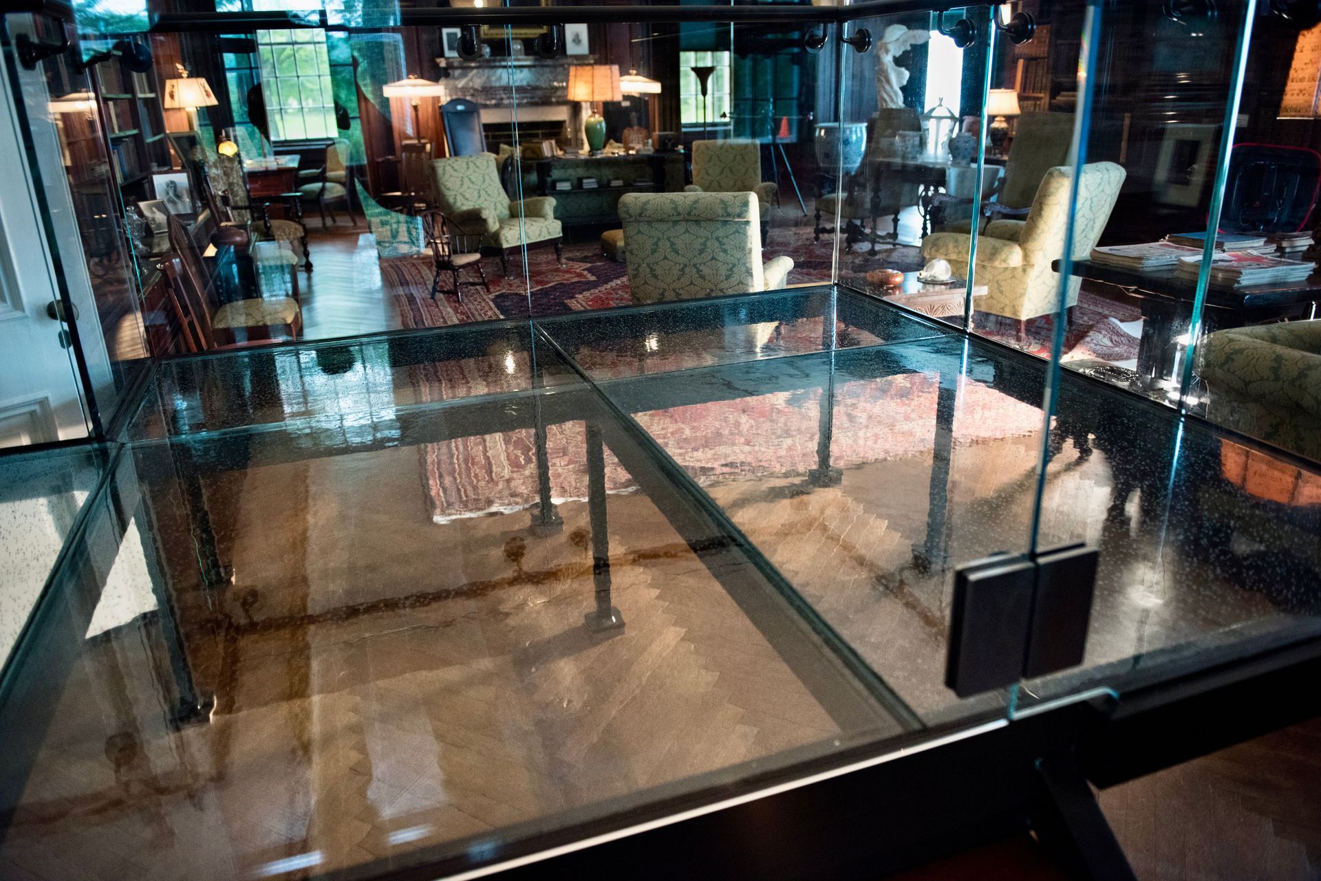 Glass floor section revealing a cozy interior with rugs, armchairs, lamps, and a fireplace.