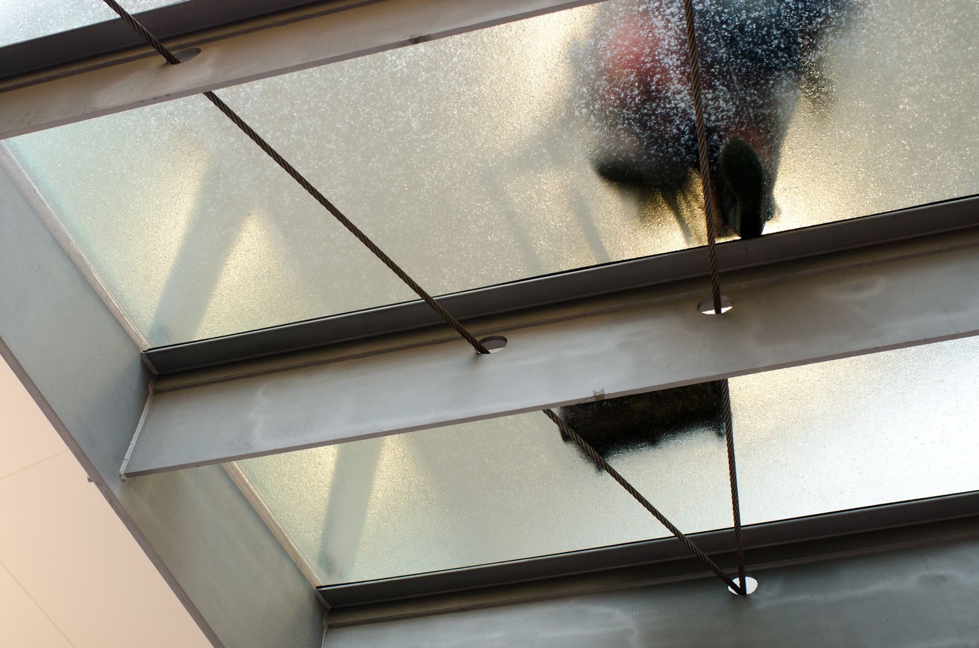 Glass ceiling paneling with person walking above, viewed from below. Metallic beams and support cables.