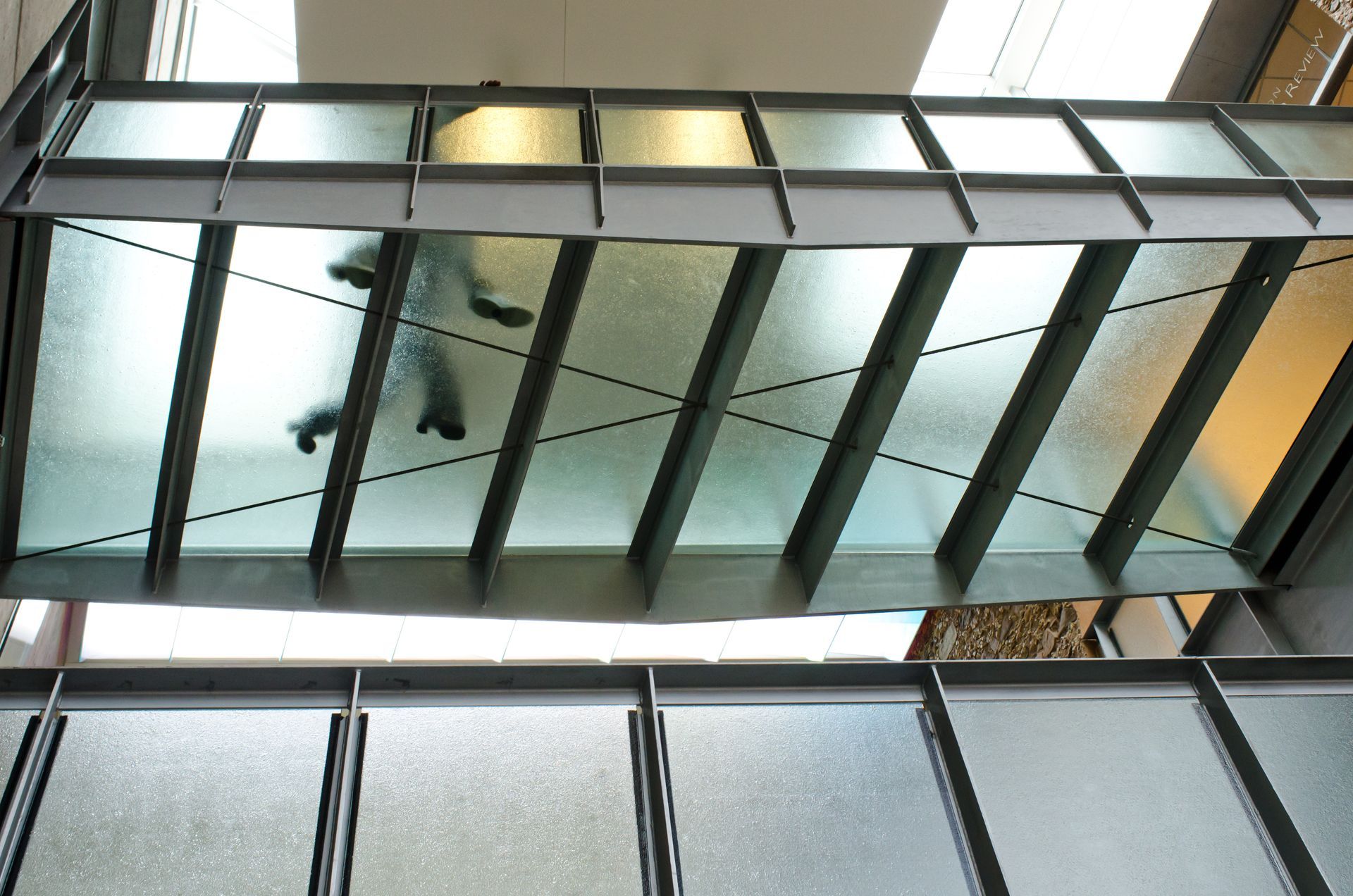 Looking up at a glass-bottomed bridge with a person walking across it. Steel beams support the structure.