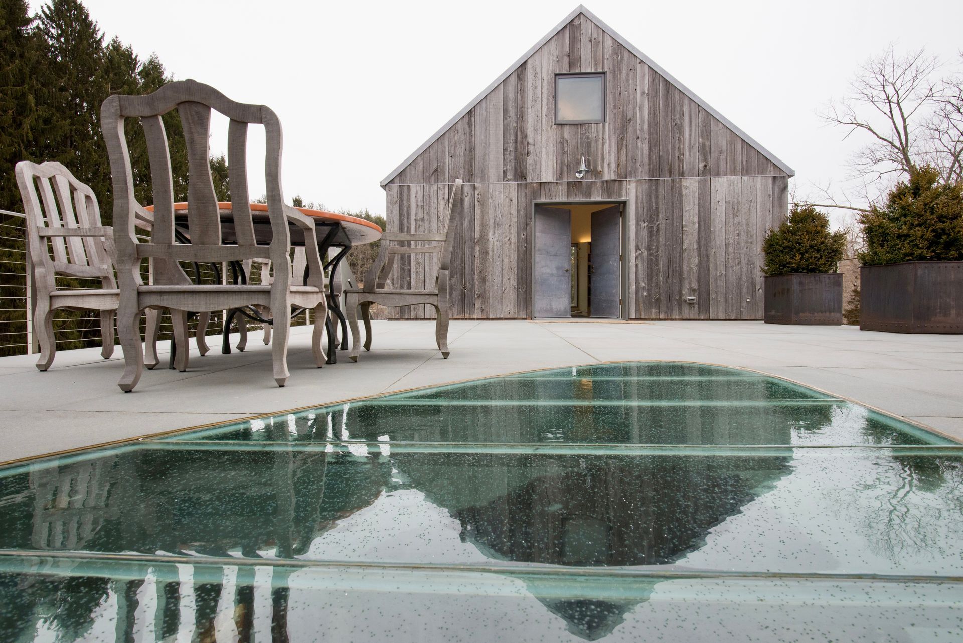 A weathered wood barn with open doors sits on a concrete patio with glass floor panels; outdoor seating is nearby.