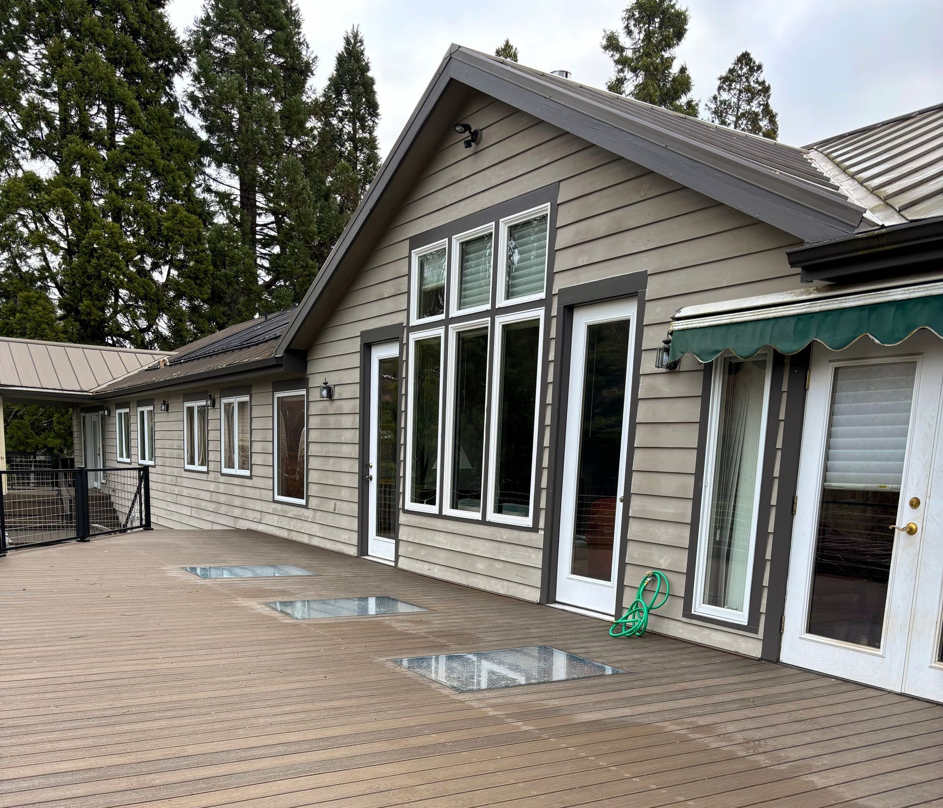 Beige house with large windows, brown trim, and a wooden deck. Trees in the background.