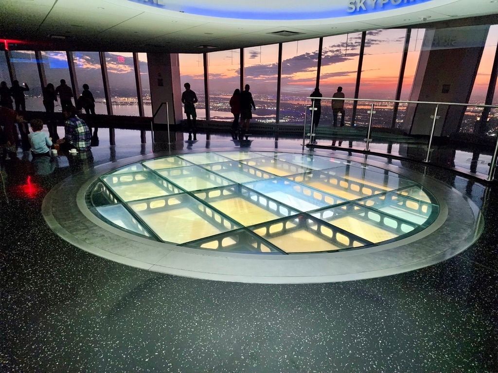 Observation deck with a glass floor, people silhouetted against a sunset view of the city.