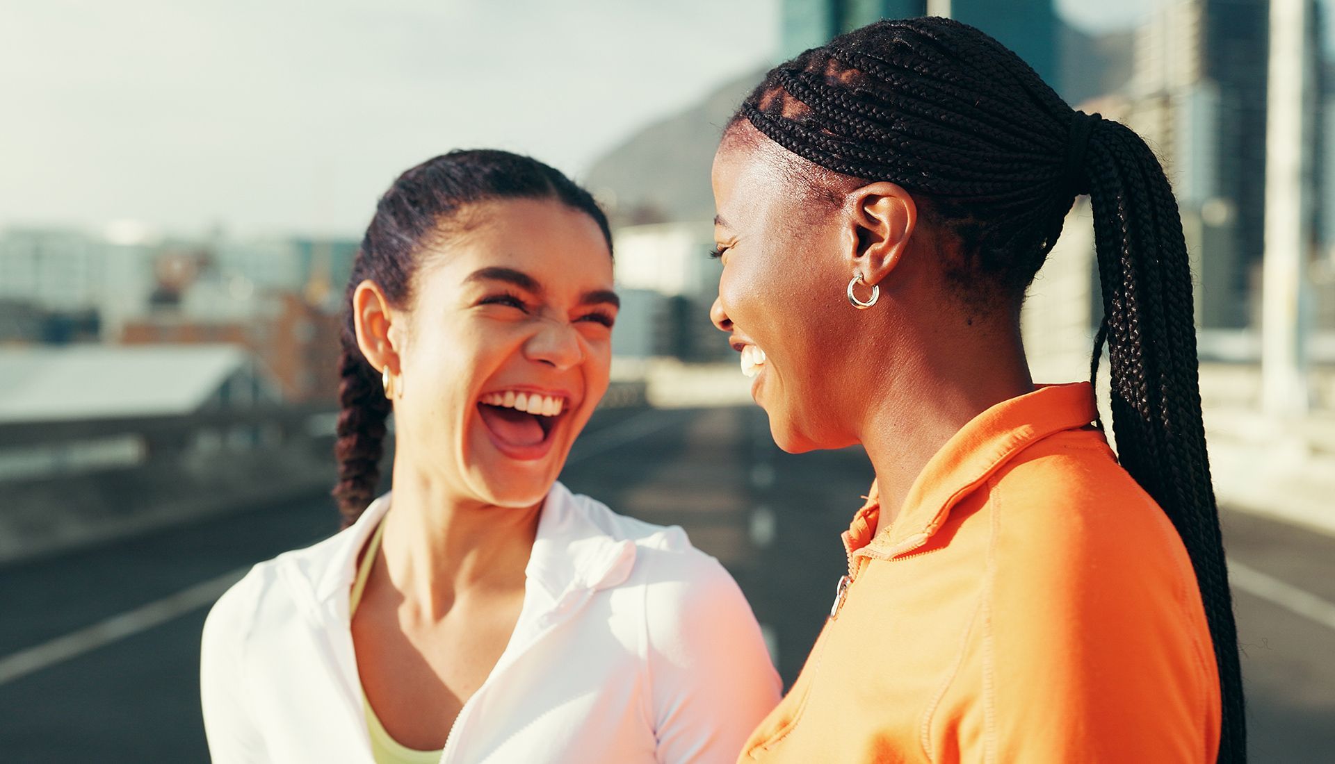 Two women laughing outdoors, one in white, the other in orange. City background.