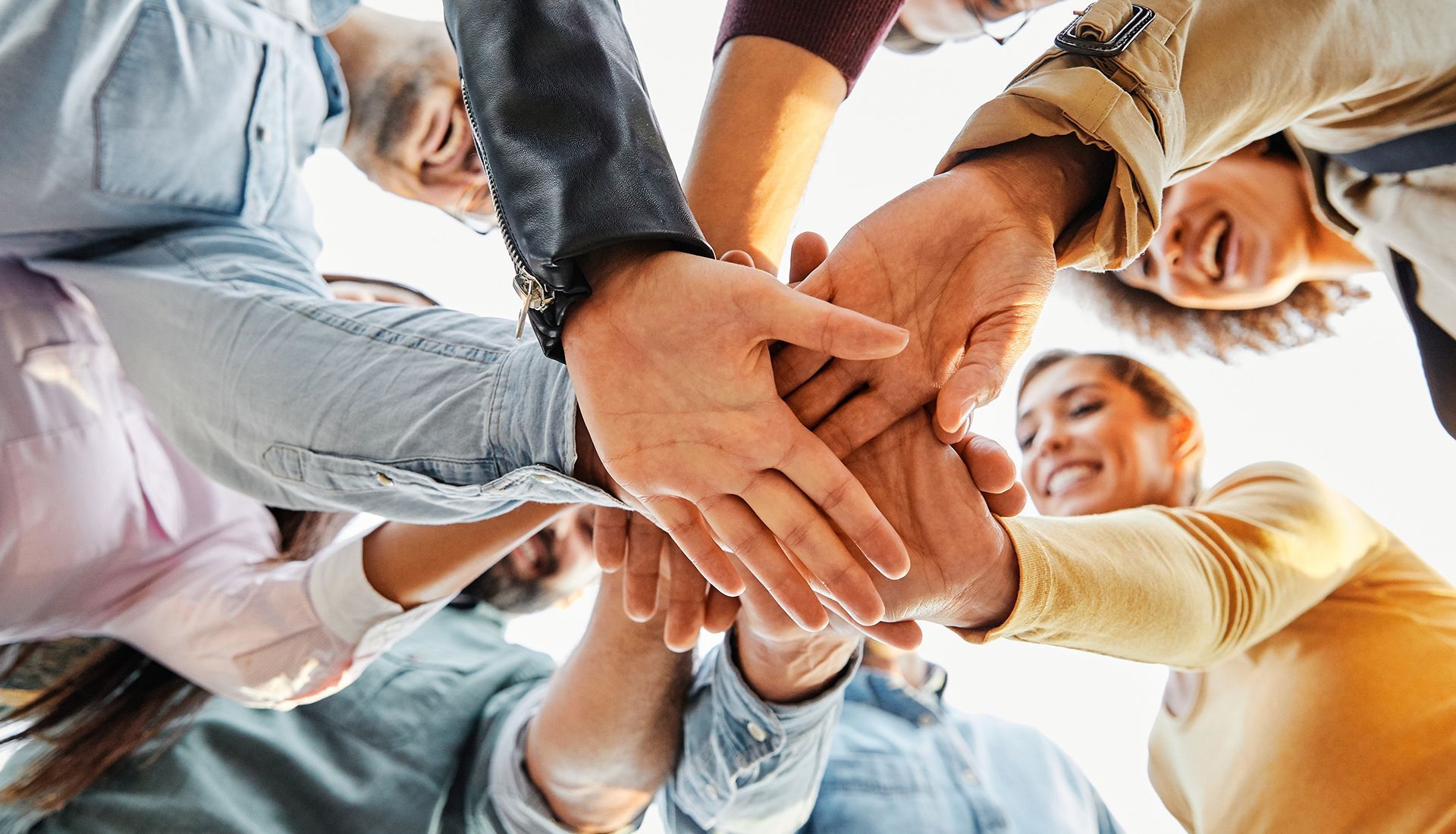 Hands joined in a huddle, smiling people, low-angle shot, sunlit.