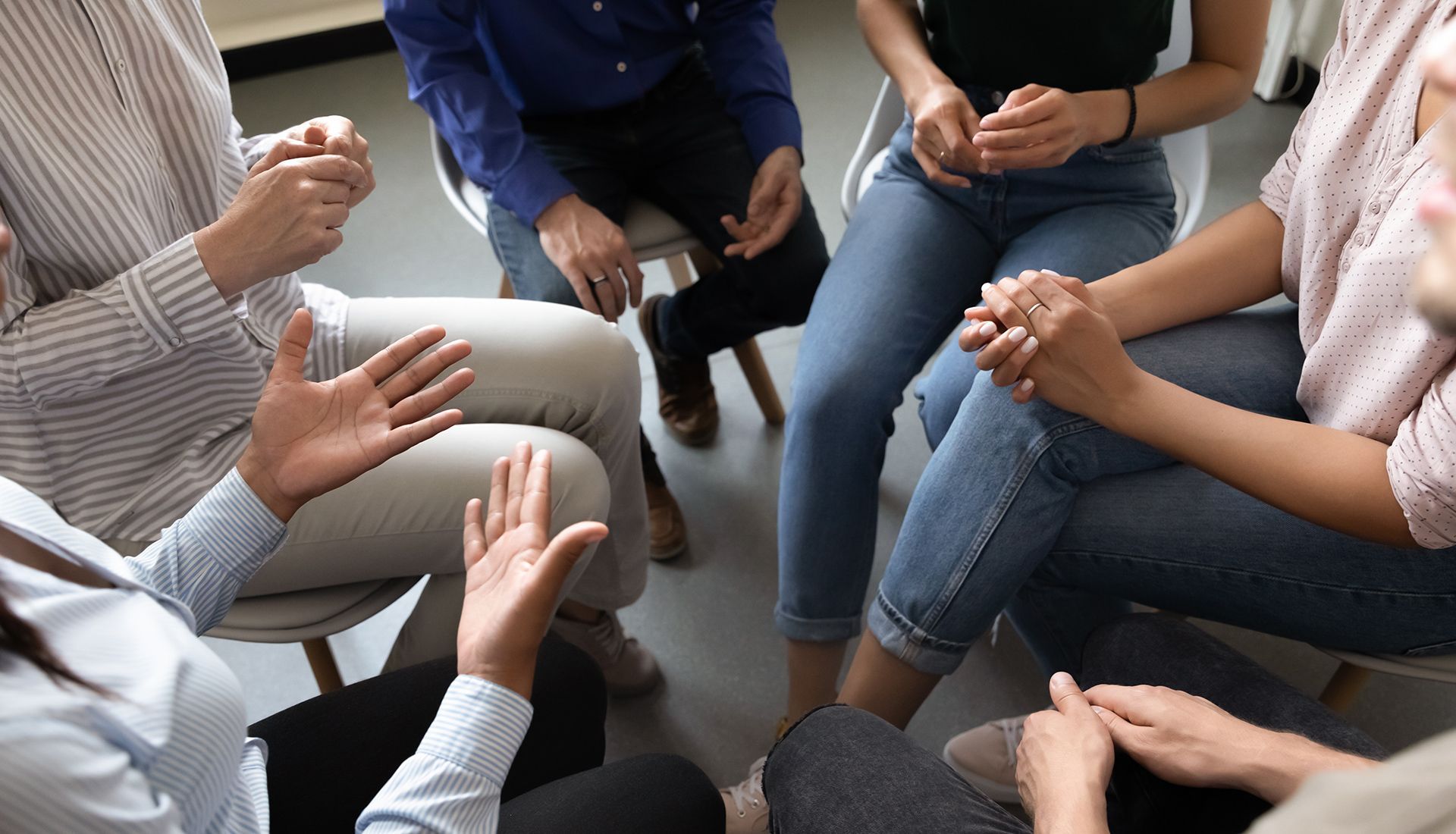 People sitting in a circle, hands clasped or gesturing, in a discussion.