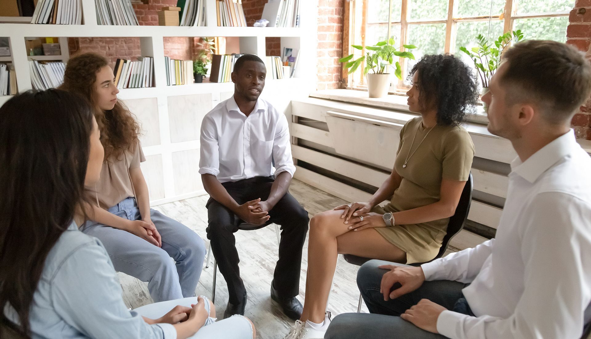 Group therapy session: Six people seated in a circle, conversing in a well-lit room.
