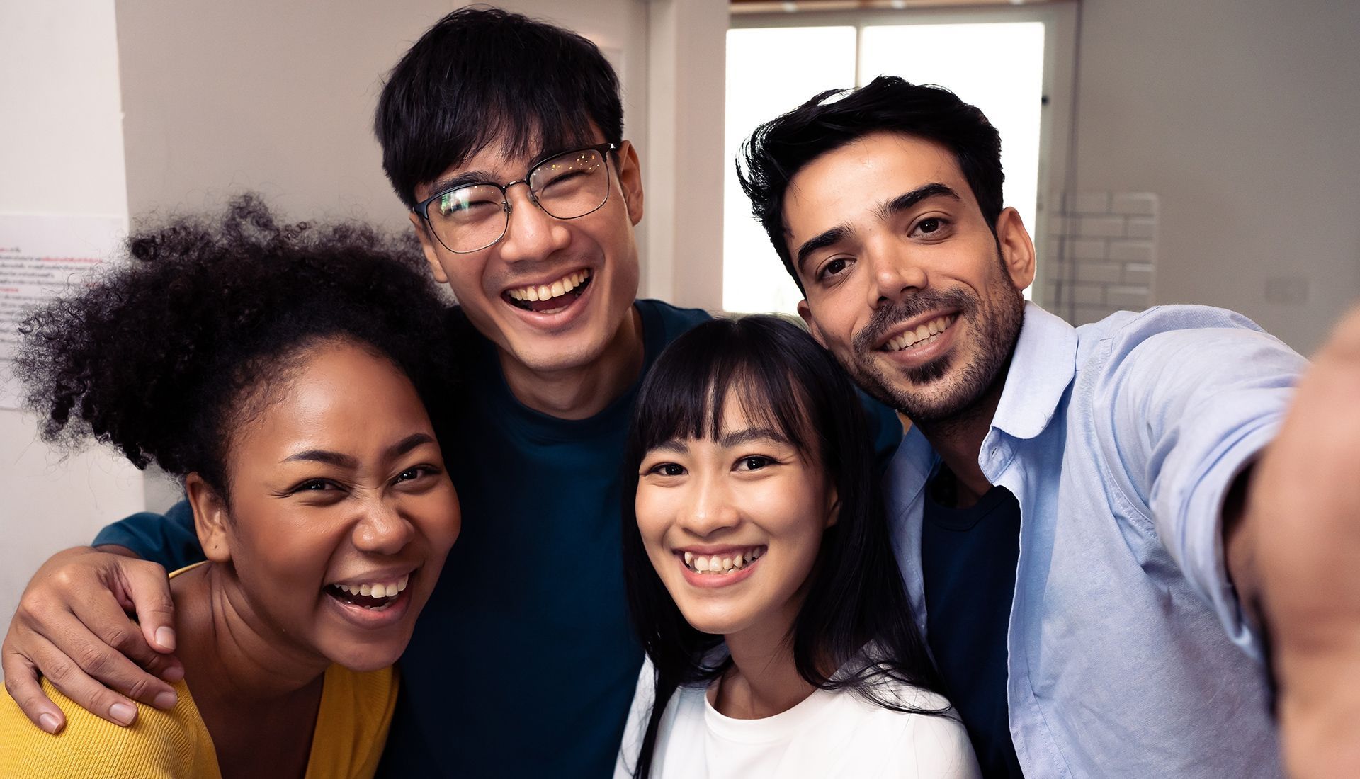 Four people smiling and taking a selfie indoors. They are of different ethnicities and are close together.