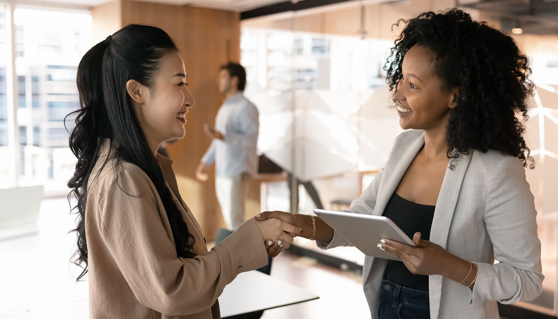 Two women shaking hands in an office, smiling. One holds a tablet. A man walks in the background.