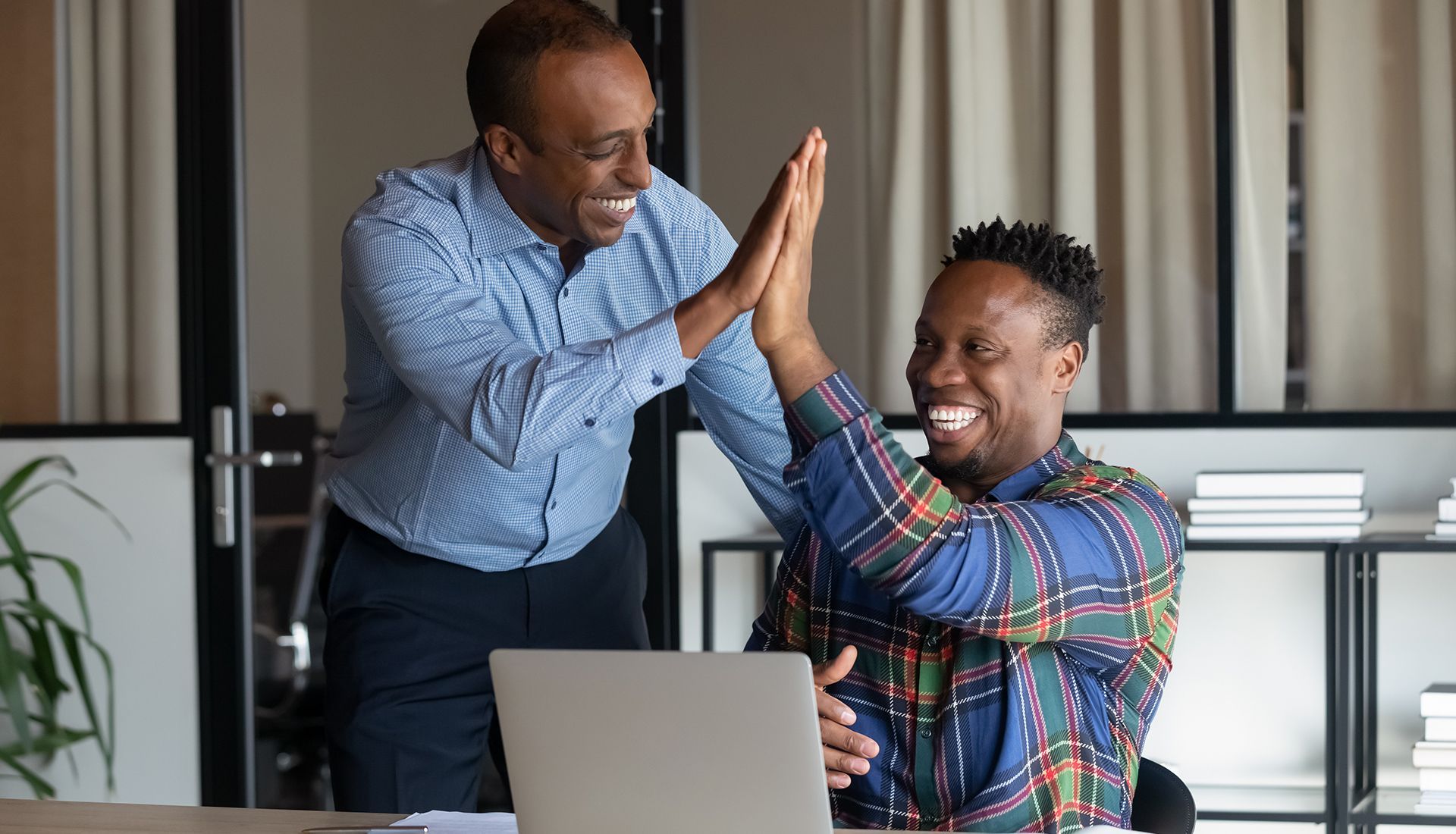 Two men high-fiving over a laptop, smiling. Office setting with natural light and a shelf.