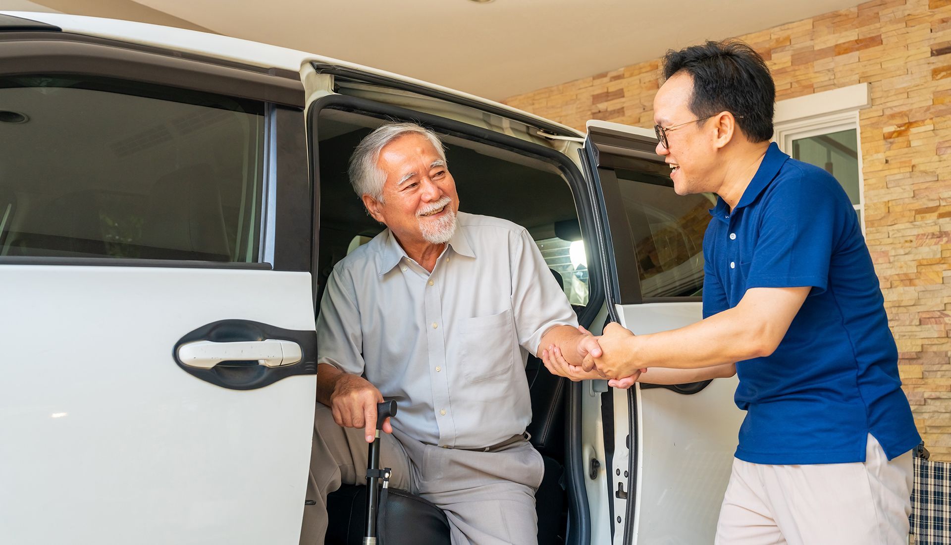 Man helping another man exit a white car. The older man uses a cane, both smile. Exterior setting.