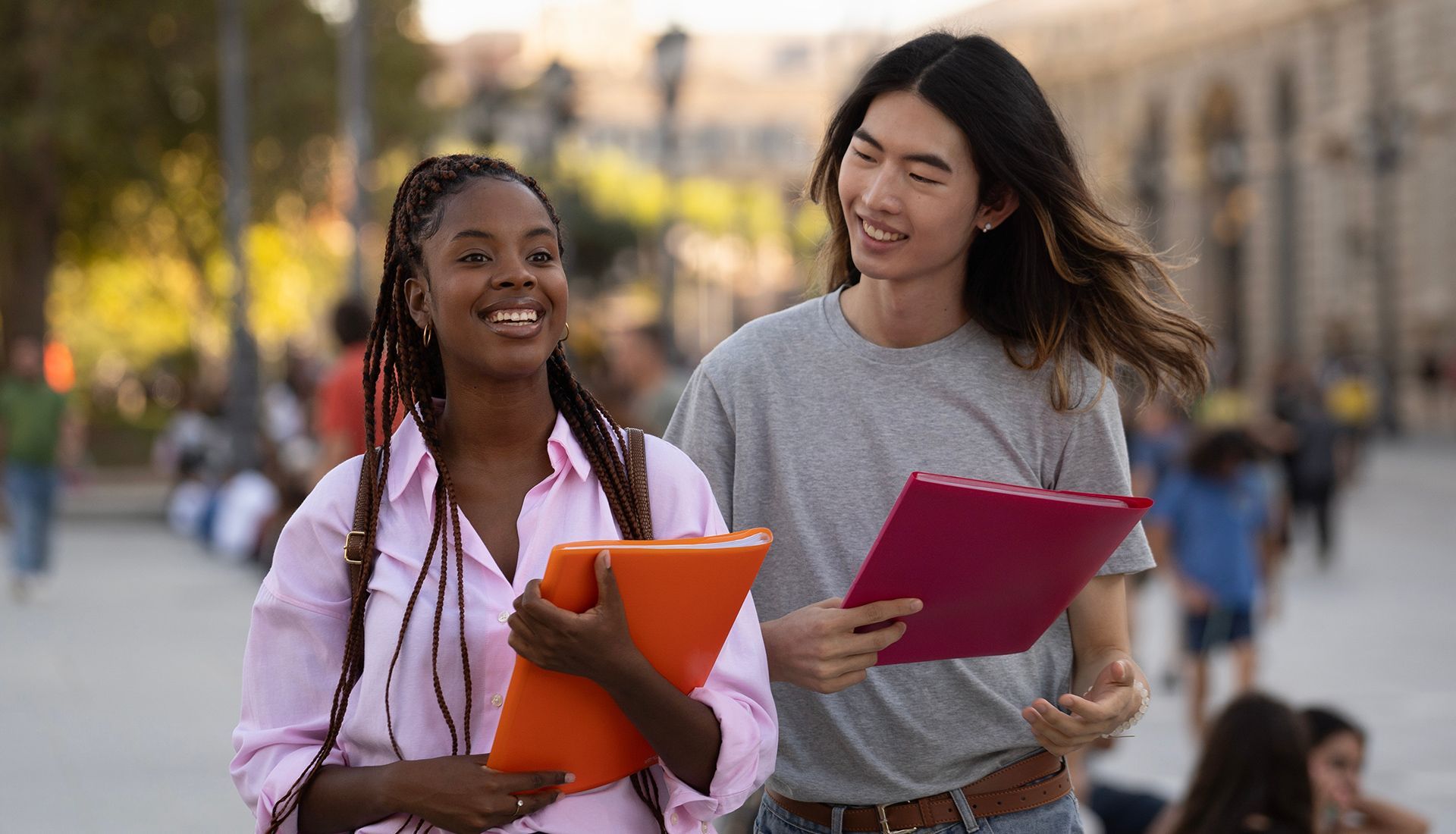 Two students walk, holding folders, smiling on a city street.