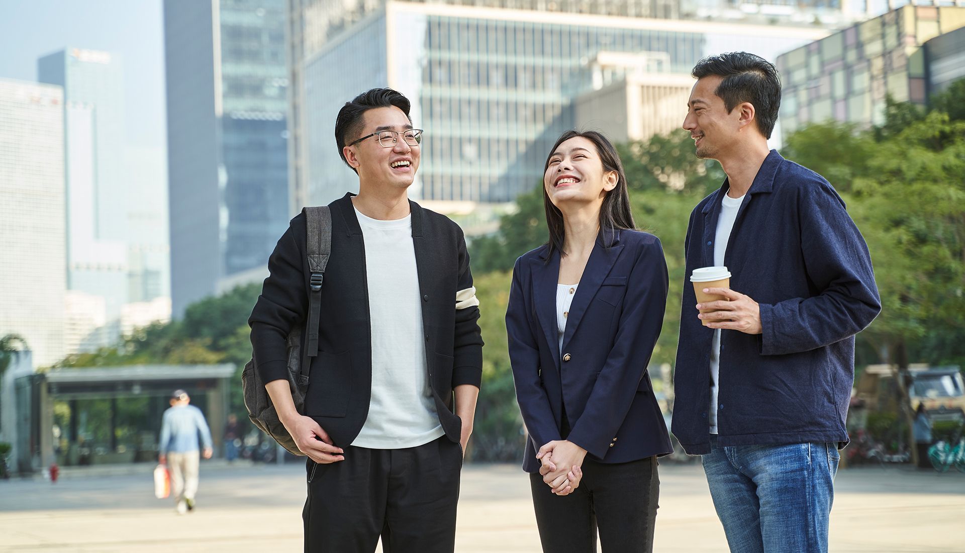 Three people smiling, talking outside, urban setting with buildings, bright daylight.