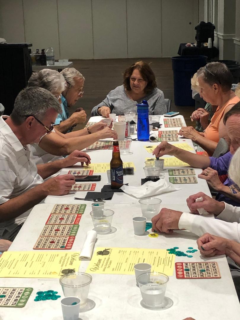 People playing bingo around a white table in a brightly lit room, using cards and markers.