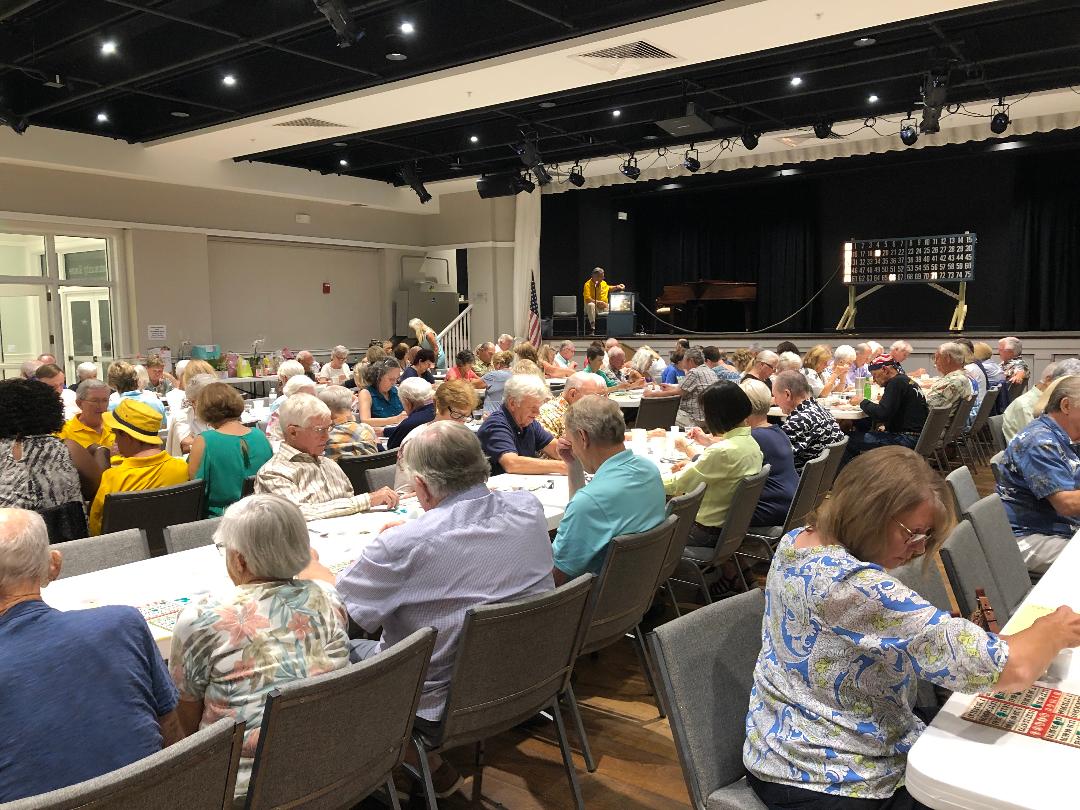 Large room filled with many people seated at tables. Stage at the back, a person on stage.