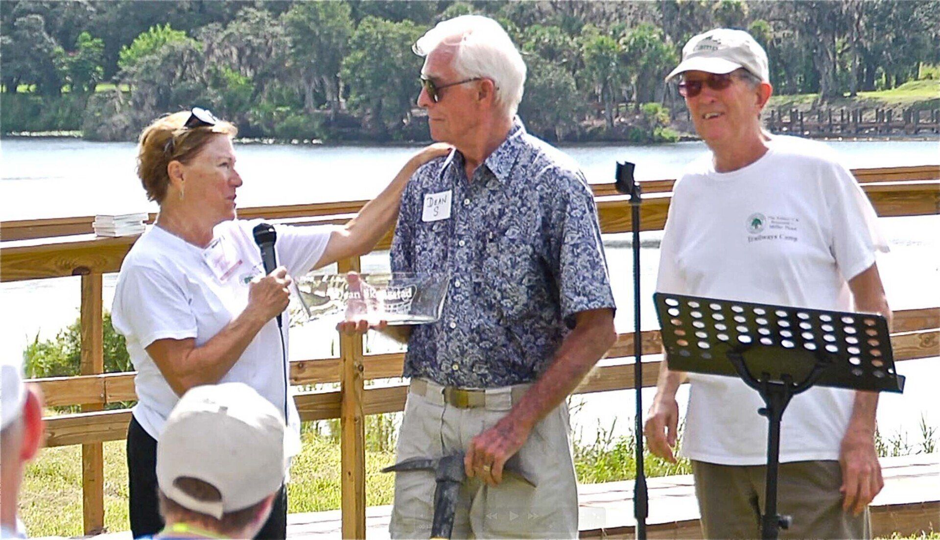 Three people at an outdoor event. A woman with a microphone, a man with sunglasses being congratulated
