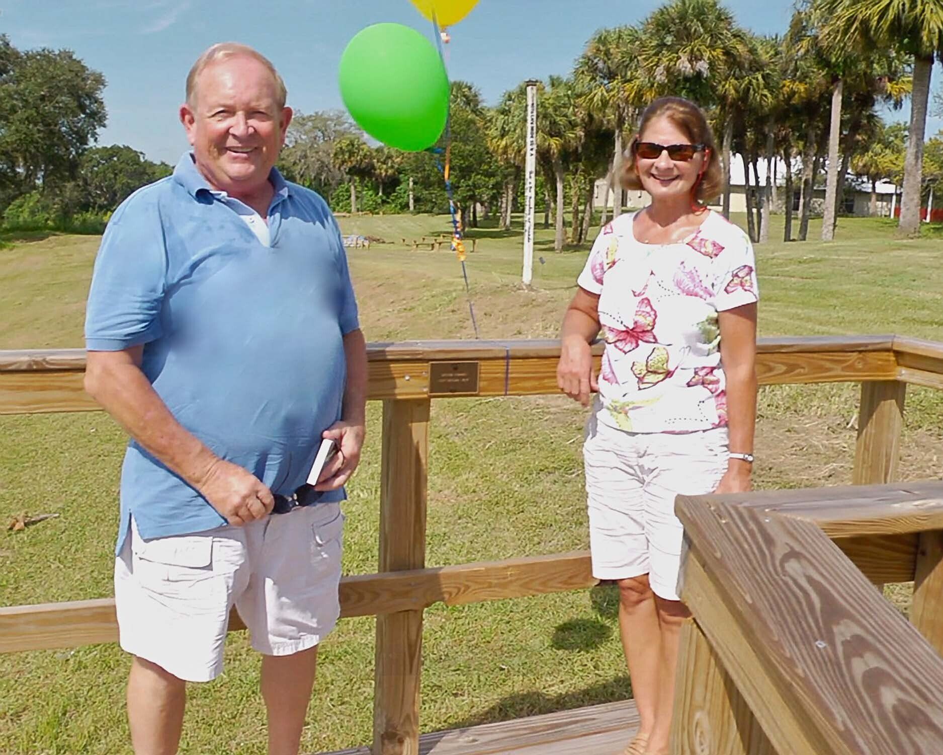 Man and woman on a wooden deck outdoors, smiling. Man in blue shirt, woman in floral top. 