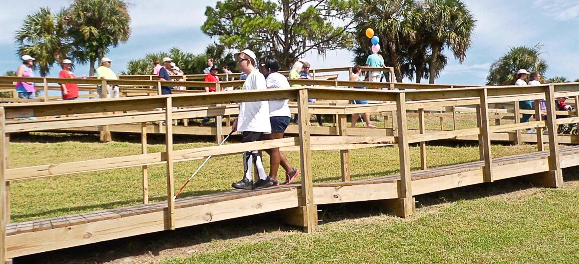 People walking on a wooden boardwalk with railings, grass in the foreground, trees and sky in the background.