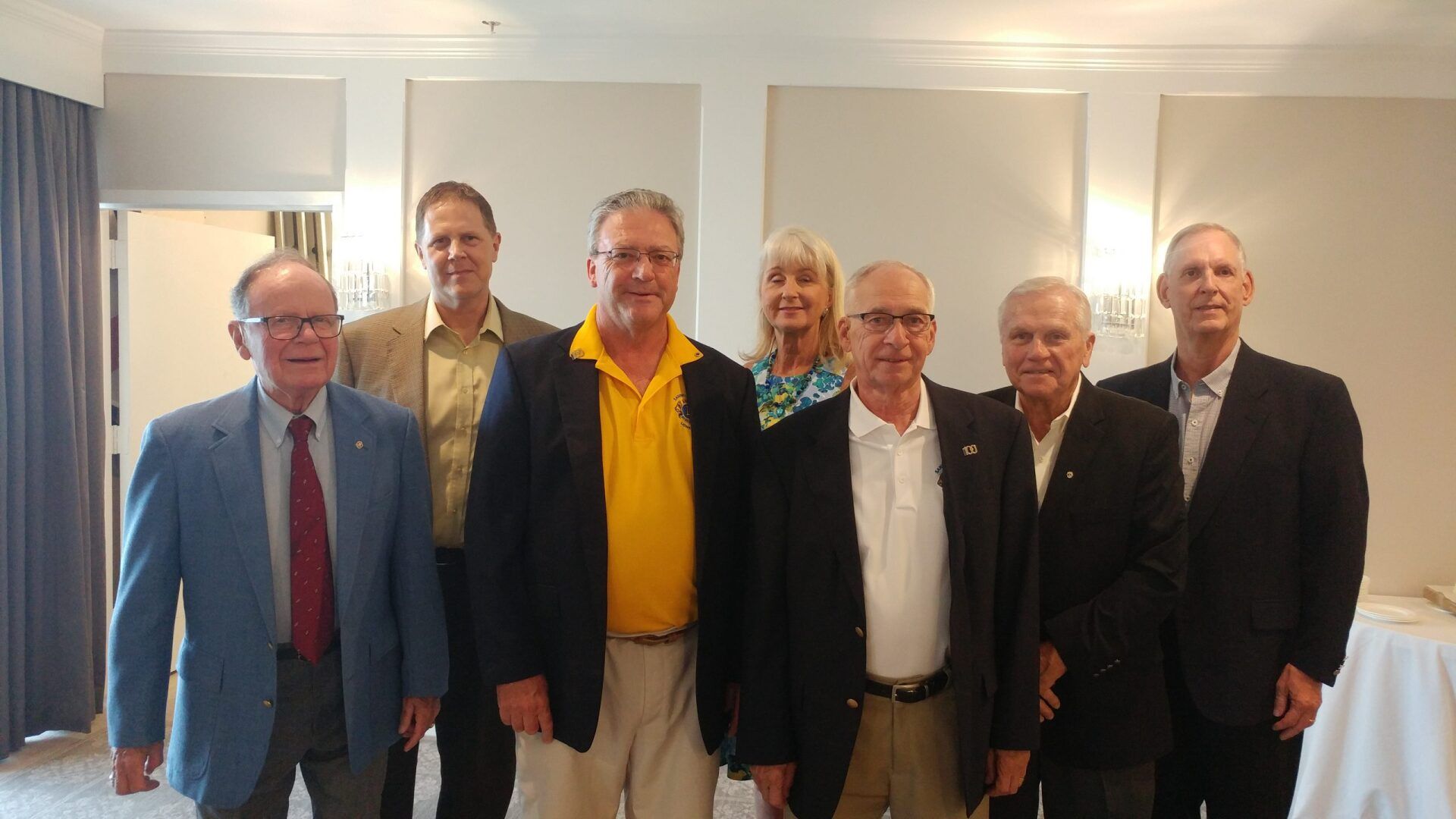 Group of eight people, mostly men, posing for a photo in an interior setting with neutral-colored walls.
