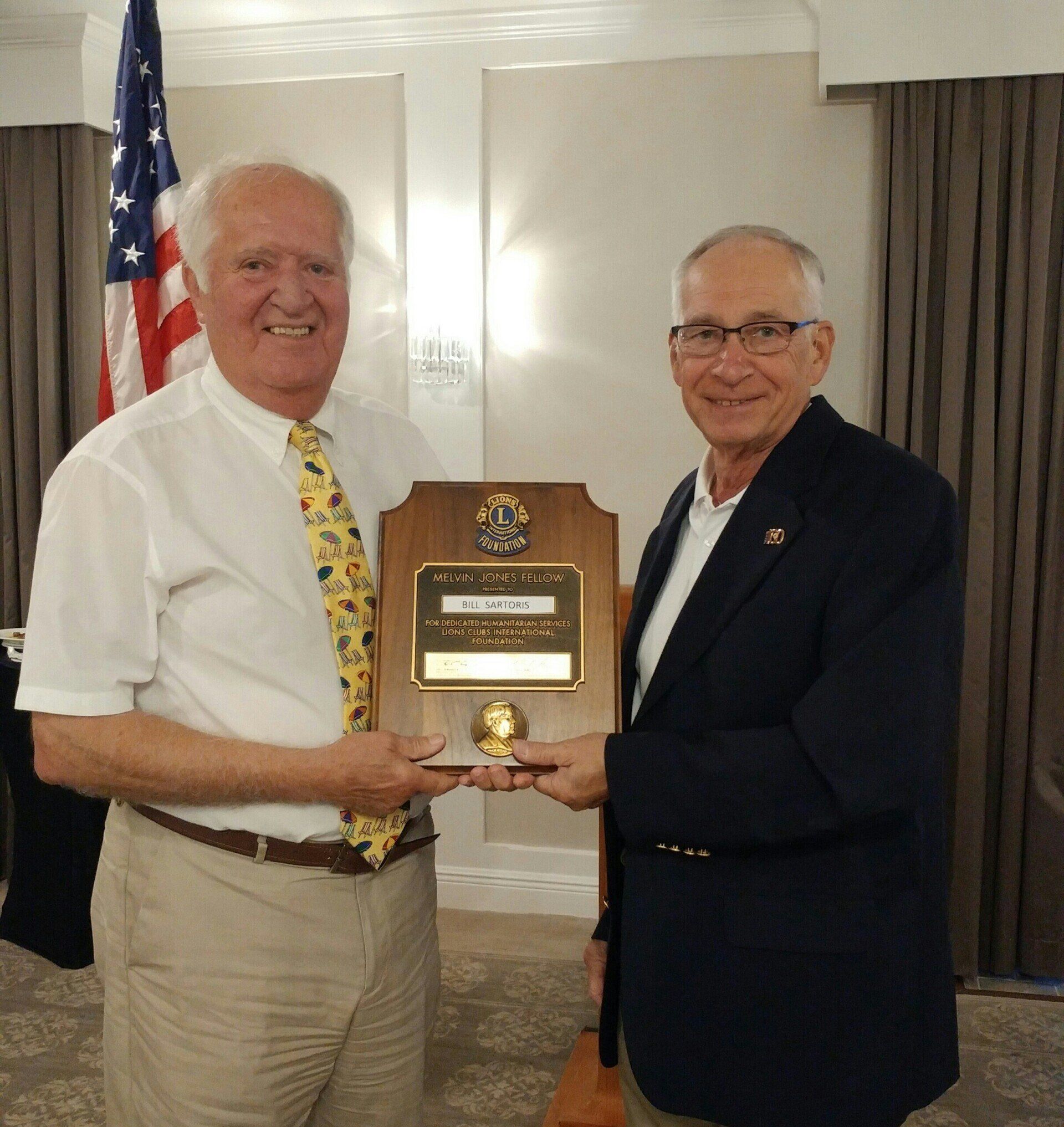 Two men holding a wooden plaque, one smiling in a yellow tie and the other in a blazer, in a room with an American flag.