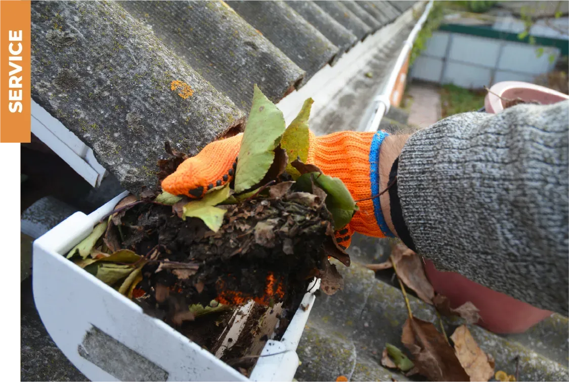 A person wearing orange gloves is cleaning a gutter with leaves