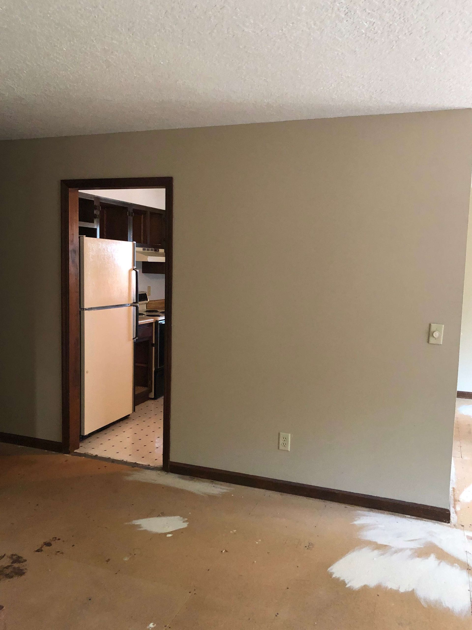 An empty room with a refrigerator and a door leading to a kitchen