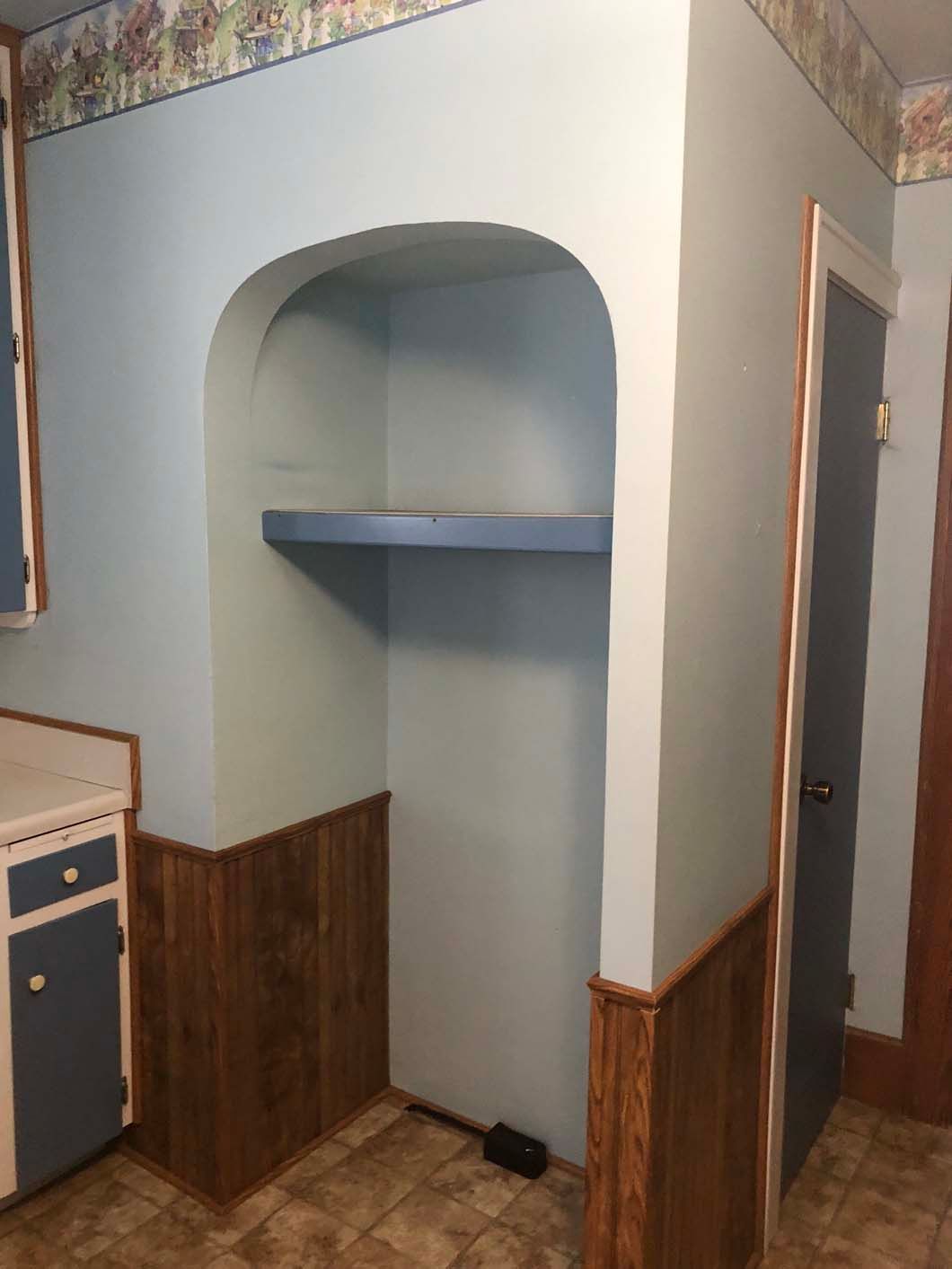 A kitchen with a shelf in the corner and a door.