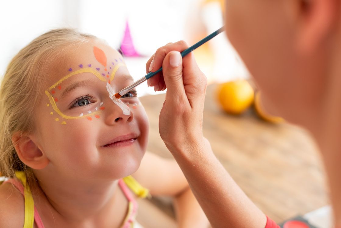 Girl getting face painted with colorful design; indoor setting.