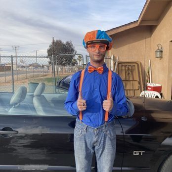 Person in orange and blue outfit, holding thumbs up, next to a black car. Sunny outdoor setting.