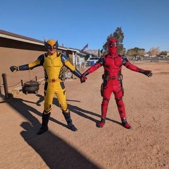 Two costumed figures, Wolverine in yellow and Deadpool in red, holding hands outdoors in a sunny, desert-like setting.