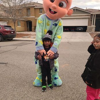 A child in a Cocomelon costume holding a smaller child on a residential street; another child smiles nearby.