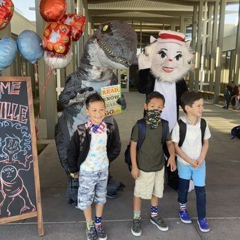 Children with a dinosaur and Cat in the Hat at a school. Balloons and a welcome sign are in the background.