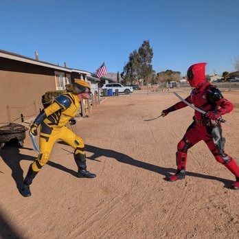 Two people in superhero costumes face off outdoors. One in yellow, one in red, wielding blades.