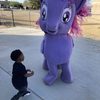 Child kneels before a large purple pony costume outdoors on a sunny day.