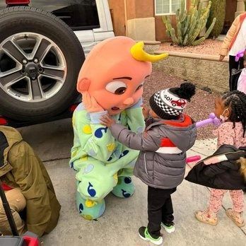 Children interact with a Cocomelon mascot in a driveway; one child touches the costume.