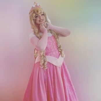 Princess Aurora in pink dress, smiling with hands near face. Pink, pastel background.