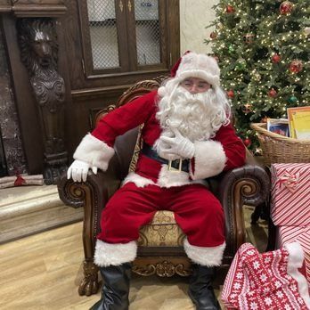 Santa Claus sits in an ornate chair, wearing a red suit, white beard, and hat, near a Christmas tree.
