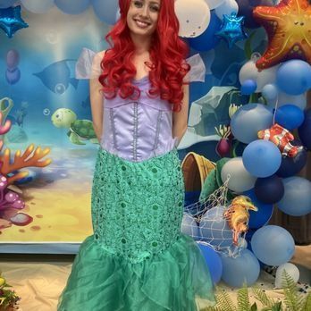 Woman in Ariel costume smiling, posing in front of a themed backdrop with balloons.