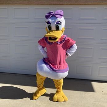 Daisy Duck mascot in pink dress, purple bow, yellow legs and feet, standing in front of a garage.
